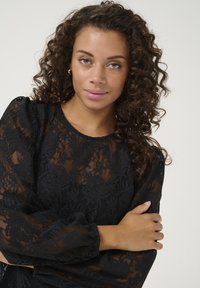 Top en dentelle noire avec des motifs floraux, manches bouffantes longues et col rond. Le tissu est transparent avec une finition texturée.