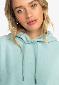 Sudadera con capucha de forro polar azul claro que presenta un cordón en la capucha, un sutil logo bordado y una textura en la tela. Acabado liso con mangas largas.