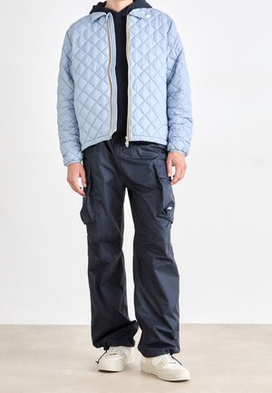 Personne portant une veste matelassée bleu clair par-dessus un sweat à capuche noir, un pantalon cargo foncé et des baskets blanches, debout devant un fond blanc uni.