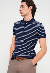 Μπλε ναυτικό πόλο με λευκές ριγέ λεπτομέρειες, κοντά μανίκια και κολάρο. Καφέ ζώνη με ορθογώνια αγκράφα. Ανοιχτό καφέ παντελόνι.