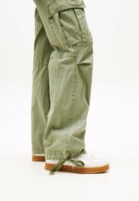 Valkoisesta kankaasta valmistetut cargo-housut, joissa on rento istuvuus, sivutaskut ja nauhakiinnitys helmassa, yhdistettynä valkoisiin lenkkareihin, joissa on vaaleanruskea pohja.