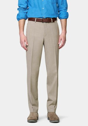Man draagt beige geplooide broek met bruine gevlochten riem, blauw overhemd met opgerolde mouwen en lichtbruine suède schoenen, rechtopstaand.