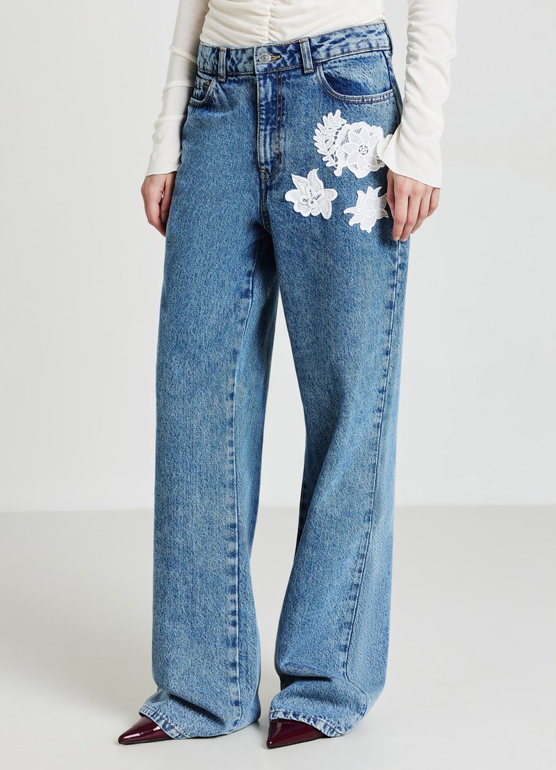 Jeans a vita alta e gamba larga in denim azzurro chiaro con ricami floreali bianchi sulla coscia sinistra. Presentano una chiusura con bottone e passanti per la cintura.
