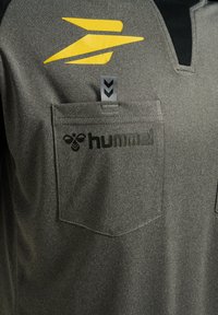Camicia grigia con texture e colletto nero, caratterizzata da un logo giallo e testo "hummel" nero sulla tasca. Include un'etichetta grigia.