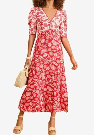 Vrouw draagt een midi-jurk met een patroon van rode en roze schelpen, met pofmouwen, houdt een geweven handtas vast en draagt beige sleehakken.