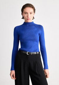 Suéter azul de gola alta com mangas longas, design justo, tecido texturizado, combinado com calças pretas de cintura alta e um cinto preto.