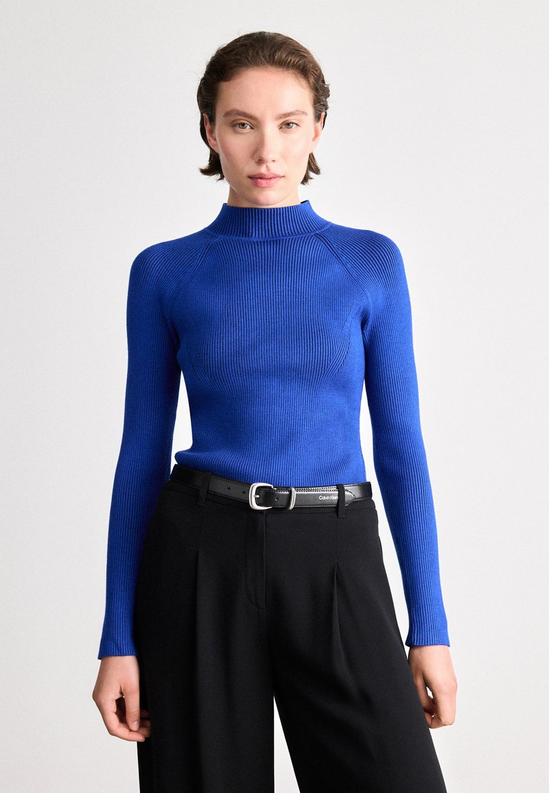 Suéter azul de gola alta com mangas longas, design justo, tecido texturizado, combinado com calças pretas de cintura alta e um cinto preto.