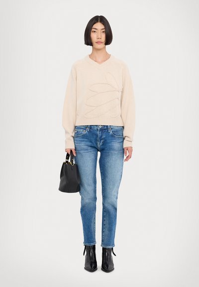 Pull beige en tricot avec décolleté en V et motif imprimé à la main, associé à un jean droit bleu et des bottines noires. Sac à main noir inclus.