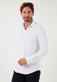 Fehér, hosszú ujjú, gombos ing klasszikus gallérral, sima felszínnel és lekerekített aljjal, diszkrét varrással és gomb részletekkel.