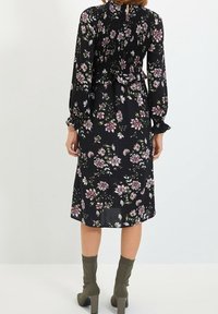Kobieta w czarnej, kwiecistej sukience midi z długimi rękawami i marszczonym bodicem, w połączeniu z oliwkowymi botkami na obcasie, zwrócona plecami.