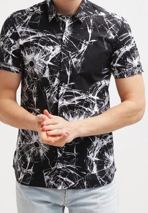 Homme portant une chemise noire à manches courtes avec un motif abstrait de verre brisé blanc, debout les mains jointes devant lui, jean délavé visible.