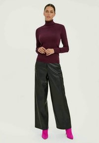 Mulher vestindo uma camisola burgundy de gola alta, calças largas de couro pretas e botas de salto rosa brilhante, de pé contra um fundo claro.