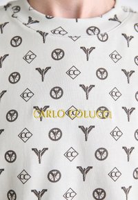 Vaaleanharmaa collegepusero, jossa on ruskeita toistuvia kuvioita, sisältäen symboleja ja "CARLO COLUCCI" -tekstiä keltaisena brodeerattuna. Teksturoitu kangas.
