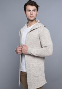Bež pleteni kardigan s kapuljačom, dugim rukavima i džepovima; lagana tekstura; složen preko bijele košulje i uparen s bež hlačama.