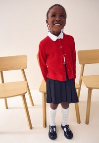 Rdeč pleten pulover s valovitim robom, bela srajca s ovratnikom, črna naborkasta krilo, bele nogavice do kolen in sijoči črni čevlji. V ozadju trije leseni stoli.