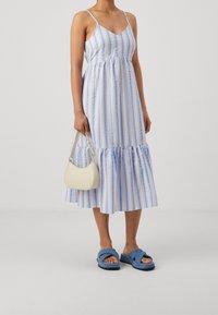 Randig midiklänning i ljusblått och krämfärgat, med tunna axelband, en samlad midja och en volangnederkant. Åtföljs av en liten krämfärgad handväska och blå sandaler.