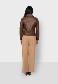 Femme aux longs cheveux bruns portant une veste en cuir marron, un pantalon large beige et des talons noirs, faisant face à un mur blanc uni, debout. 