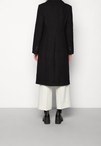 Abrigo de lana negro con una silueta estructurada, solapas anchas y puños abotonados. Llevado sobre pantalones de pierna ancha blancos y botas de tacón negras.