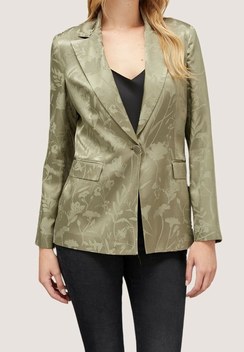 Femme portant un blazer vert clair à motif floral sur un haut noir et un pantalon sombre, debout les bras détendus le long du corps.