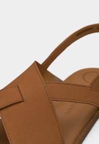 Sandalia de cuero marrón con tiras anchas cruzadas y una correa trasera abierta para soporte del talón.