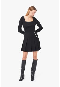 Schwarzes Kleid mit langen Ärmeln und rechteckigem Ausschnitt; hat einen plissierten Rock und drei silberne Knöpfe an der Taille. Getragen mit schwarzen knielangen Stiefeln.