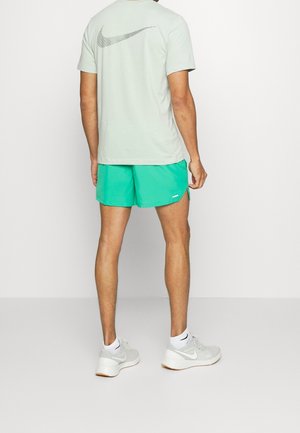 Mies, jolla on päällään vaaleanharmaa Nike-t-paita, jossa suuri logo takana, kirkkaanvihreät shortsit, valkoiset sukat ja valkoiset Nike-juoksukengät, seisoo yksivärisen taustan edessä.