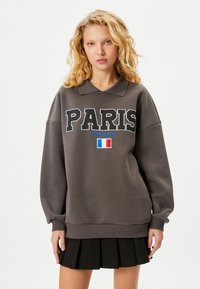 Szary sweatshirt z kołnierzykiem polo, z dużym napisem "PARIS", "Francja" w kolorze niebieskim oraz graficznym przedstawieniem flagi Francji. Długie, luźne rękawy z prążkowanymi mankietami.