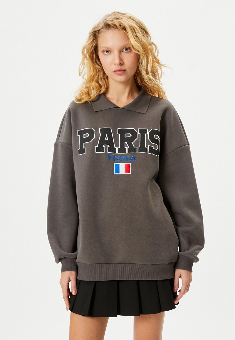 Szary sweatshirt z kołnierzykiem polo, z dużym napisem "PARIS", "Francja" w kolorze niebieskim oraz graficznym przedstawieniem flagi Francji. Długie, luźne rękawy z prążkowanymi mankietami.