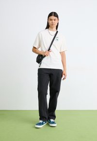 Hvid kortærmet T-shirt med logopatch, parret med sorte straight-leg jeans og teal sneakers med hvide soles. Lille sort crossbody taske.