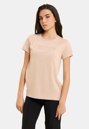 Giovane donna con lunghi capelli scuri che indossa una maglietta beige con il logo "FREDDY" e pantaloni neri, posa davanti a uno sfondo bianco uniforme.