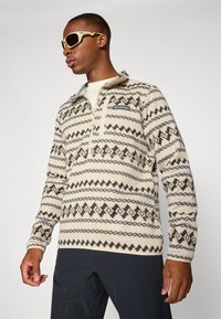 Pull en polaire crème avec des motifs géométriques noirs et beiges. Doté d'un col zippé mi-hauteur et d'un logo sur la poche poitrine. Porté avec des lunettes de soleil.
