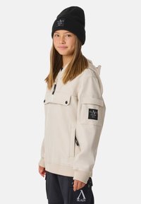 Giacca beige con cappuccio, chiusura frontale con zip, tasche laterali e patch con logo sulla manica; abbinata a un berretto a maglia nero. Texture liscia, vestibilità comoda.