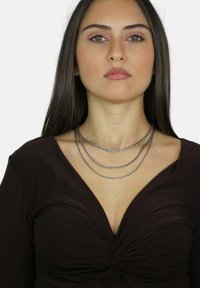 Trzy warstwowe srebrne łańcuchy, z skręconymi ogniwami, noszone na ciemno brązowej koszulce z głębokim dekoltem w kształcie V, zdobione węzłem na środku.