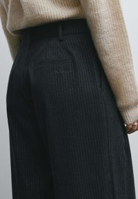 Pantalon à rayures fines sombres avec une coupe ajustée, doté de poches arrière et d'un logo brodé. Porté avec un pull en maille crème.