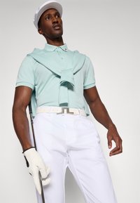 Polo de cor azul-clara com colarinho, cinto branco e calças brancas. Um taco de golfe é segurado com uma mão, e uma luva branca é usada na outra.