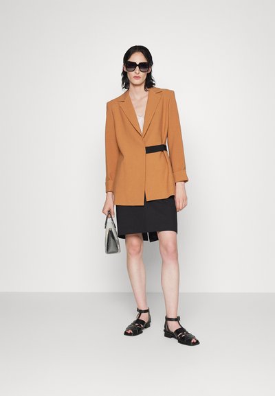 Blazer marron au design structuré, ceinture noire, associé à une jupe noire et des sandales noires à lanières. Les accessoires comprennent un petit sac à main gris.