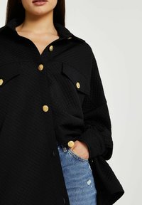 Svart kviltad skjortjacka med krage, som har guldtonade knappar och två bröstfickor. Modellens hand vilar i denimbukser.