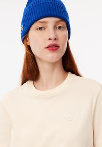 Camiseta de color crema con cuello redondo y detalle de logo en relieve. El modelo lleva un gorro de punto acanalado de color azul brillante.