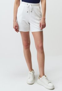 Pantaloni corti in cotone bianco con vita regolabile, caratterizzati da accessori in oro, abbinati a sneakers bianche con un accentuazione beige sul tallone.