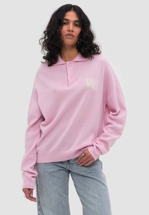 Vrouw met krullend donker haar, gekleed in een lichtroze lange-mouwen trui met kraag en blauwe spijkerbroek, poserend met hand op heup.