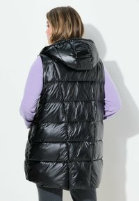 Schwarze, wattierte Weste mit Kapuze, die ein gestepptes Design und eine glänzende Oberfläche aufweist. Getragen über einem lila Langarmoberteil.