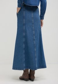 Femme portant une longue jupe en denim bleu et une veste en denim assortie, se tenant les mains dans les poches et portant des bottes cheville marron.