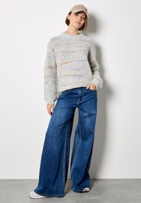 Maglione grigio a maglia texture con righe multicolori, abbinato a jeans a gamba larga blu e un cappellino beige, su uno sfondo neutro.