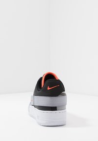 Svart och grå Nike-sneaker med ett texturerat rem, orange detaljer och en tjock vit sula. Synlig branding på hälen och sidan.