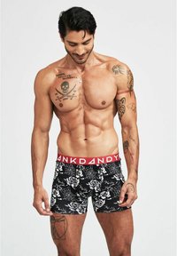 Svarta boxershorts med ett mönster av vit ros och spindelnät, med en röd midja märkt "DANDY." Slät tygkvalitet, figurnära design.