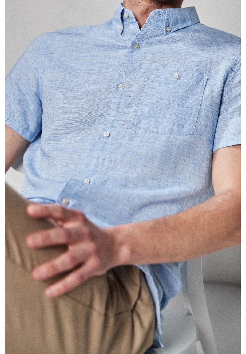 Chemise bleu clair à manches courtes avec poche, confectionnée en tissu texturé, dotée de surpiqûres contrastantes et de boutons blancs.