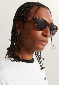 Kočičí slunční brýle v želvovinovém vzoru s tmavými skly, obdélníkový rám a hladký povrch. Model má zapletené vlasy, na sobě má světle šedou košili s černými akcenty.