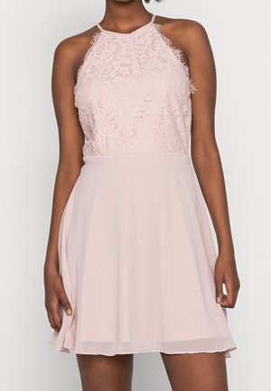 Robe rose pâle sans manches avec un décolleté halter en dentelle et une jupe fluide se terminant au-dessus des genoux, portée par une personne à la peau foncée.