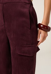 Bordeaux corduroy broek met verticale strepen, voorzien van zijzakken en een relaxte pasvorm. Een glanzende bordeaux armband siert de pols.