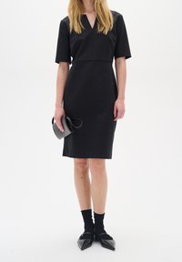 Černé šaty krátkého rukávu s výstřihem do V, přiléhavý střih, délka po kolena. Spárováno s černými balerínami s špičatou špičkou a strukturovanou černou kabelkou.